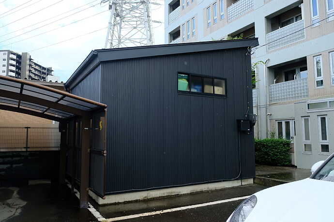 中央の黒い建物の向かって右端にメーターが見える。画面右上に見えるケーブルが電柱からの電線。画面左の屋根付き駐車場の右端に、テスラのウォール・コネクターがある。