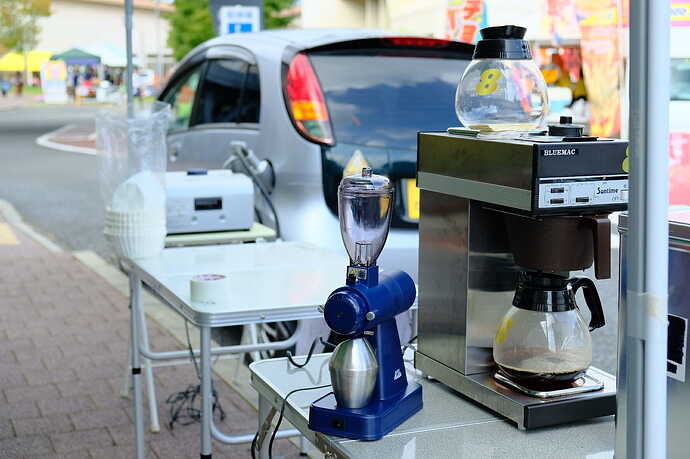 後方のi-MiEV Mタイプからの電力で、、手前のコーヒーミルと珈琲マシンを動かして珈琲を淹れているところ。MiEV Power BOX（奥のテーブル上の銀色の箱）を使えばCHAdeMOソケット経由でAC100Vが1,500Wまで出せるので、電子レンジやホットプレートも充分動かせる。富士河口湖町でのイベントにて。