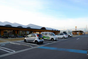 福島県北部に位置する国見町（くにみまち）にある「道の駅 国見あつかしの郷」。アイミーブの目の前は花屋さん、右奥にコンビニ、右の屋根付きの部分に車椅子駐車スペースと急速充電器がある。キャンピングカーを含め、車中泊のクルマが8台ほどいた。